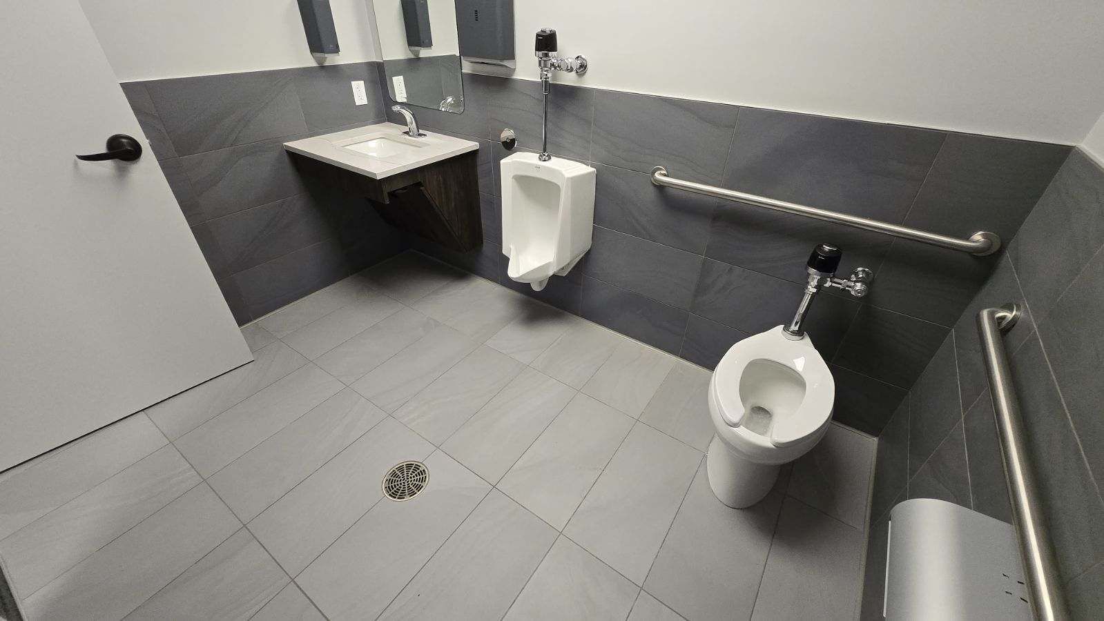 A public restroom with a sink, urinal, and toilet, featuring gray tile and grab bars for accessibility.