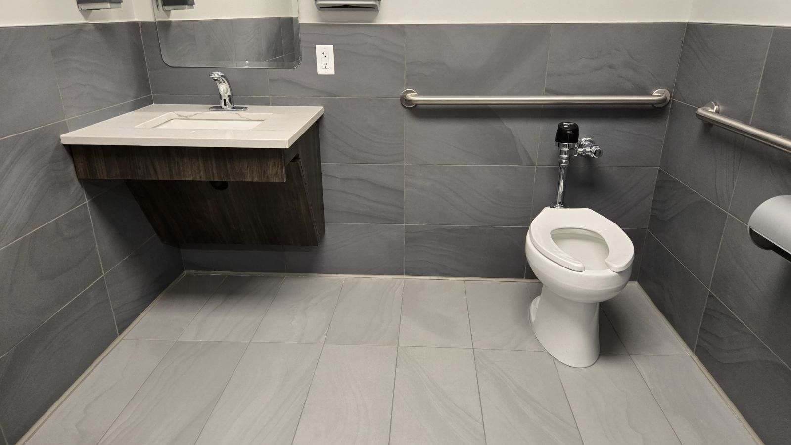 Accessible public restroom with a sink, toilet, and grab bars. Gray tiles on walls and floor.