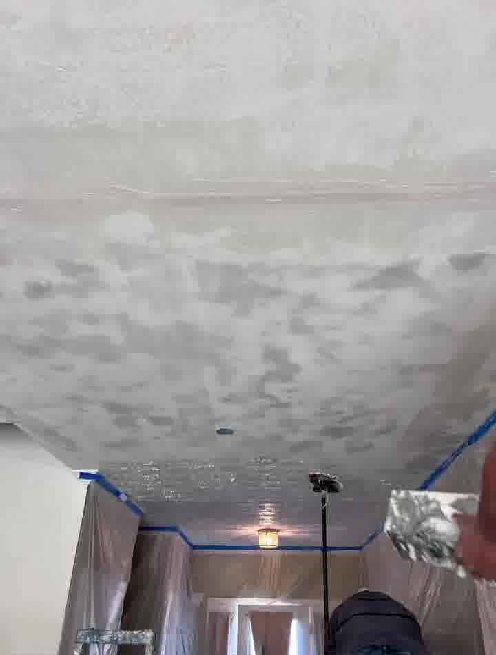 Person applying texture to a ceiling with a trowel. White, gray, and blue materials in a hallway.