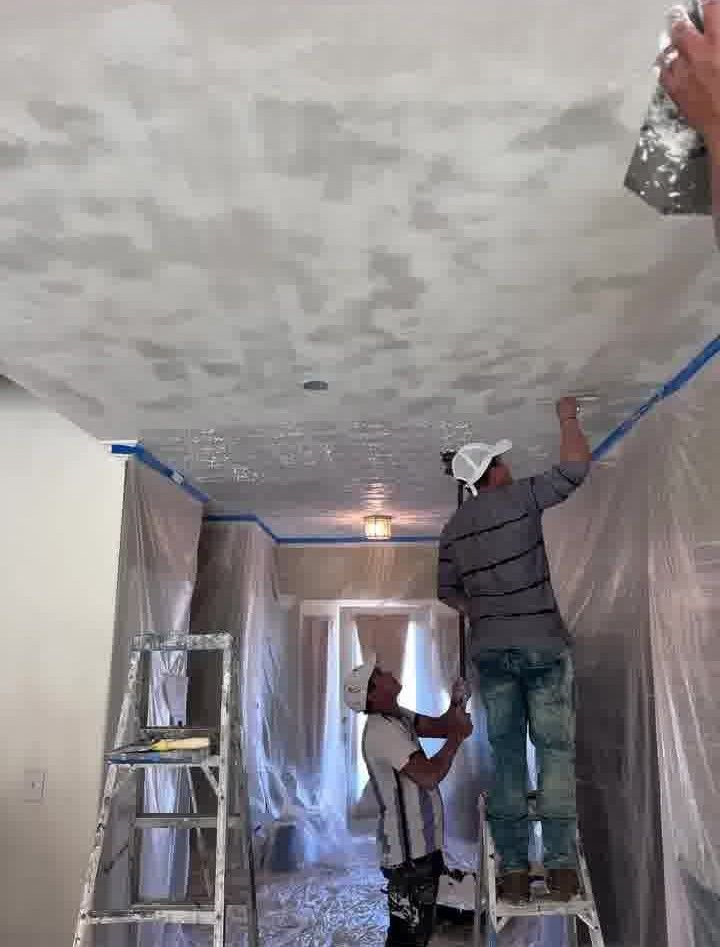 Two workers applying plaster to a ceiling. Blue tape protects edges; plastic covers walls.