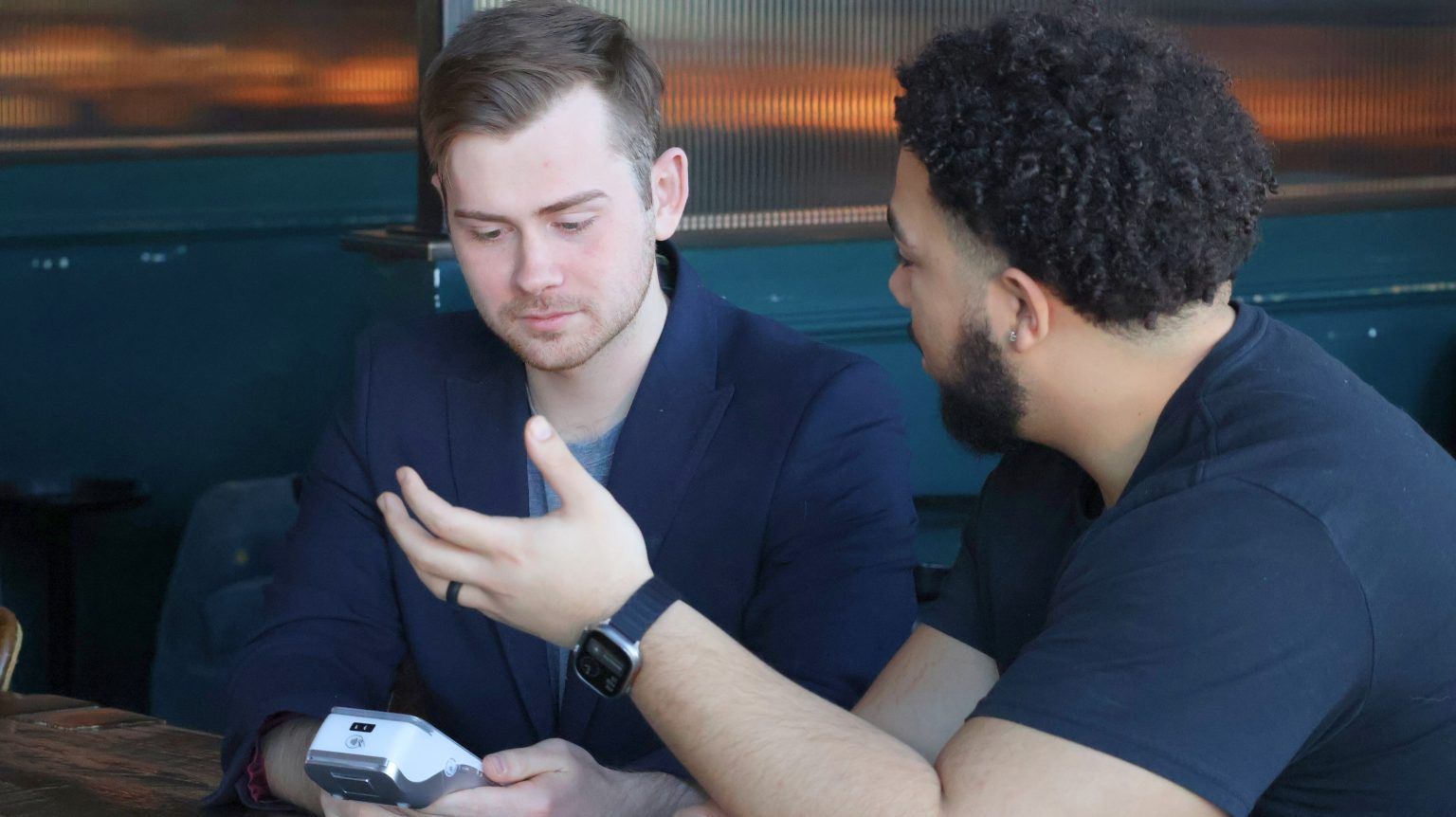 Two men are sitting at a table looking at a cell phone.