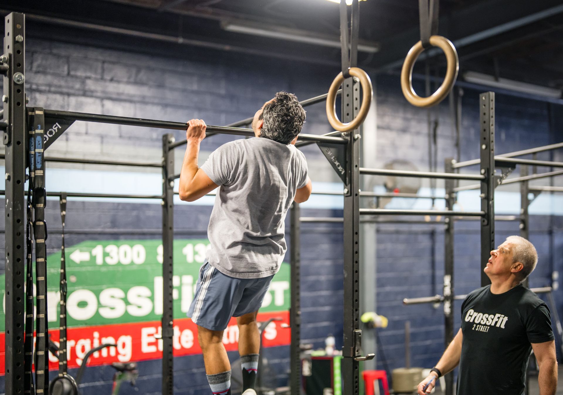 A man is doing a pull up on a bar in a gym while another man watches.