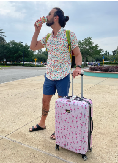A man drinking water while standing next to a pink flamingo suitcase