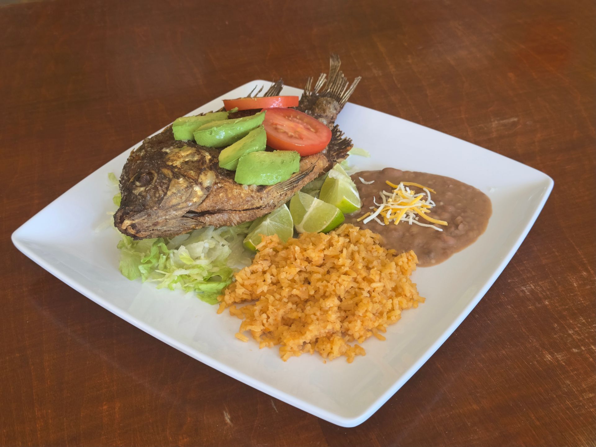 Fried fish with avocado, tomato, rice, beans, and lime on a white plate.