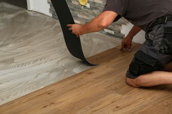 Person installing wood-look flooring, peeling back a black adhesive strip in a room with a stone wall.