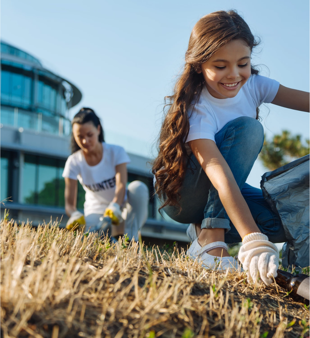 A little girl is kneeling down in the grass and picking up trash.