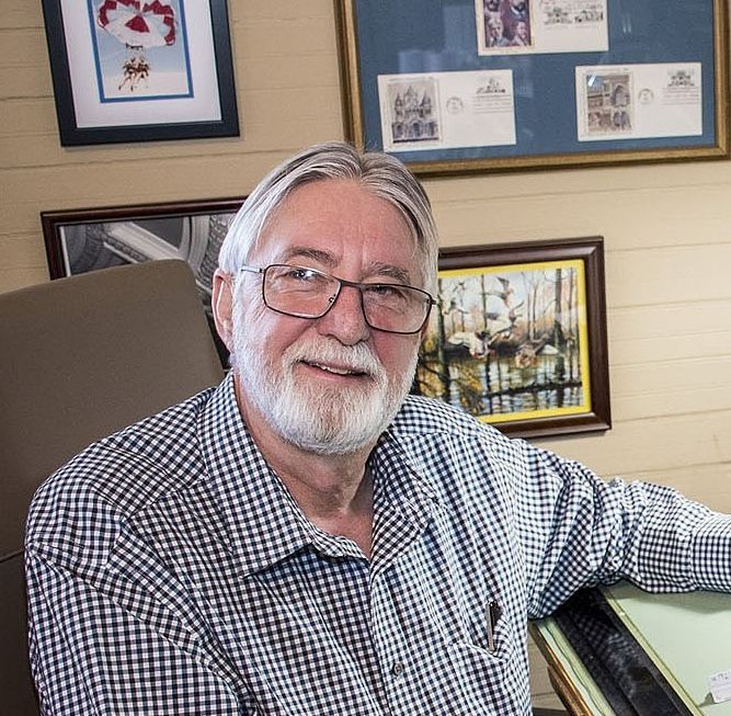 Bearded man in overalls, cap and glasses tending a garden, smiling.