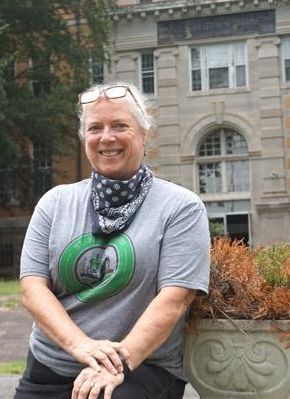Woman in front of a building, smiling. She wears glasses, a bandana, and a green logo shirt.
