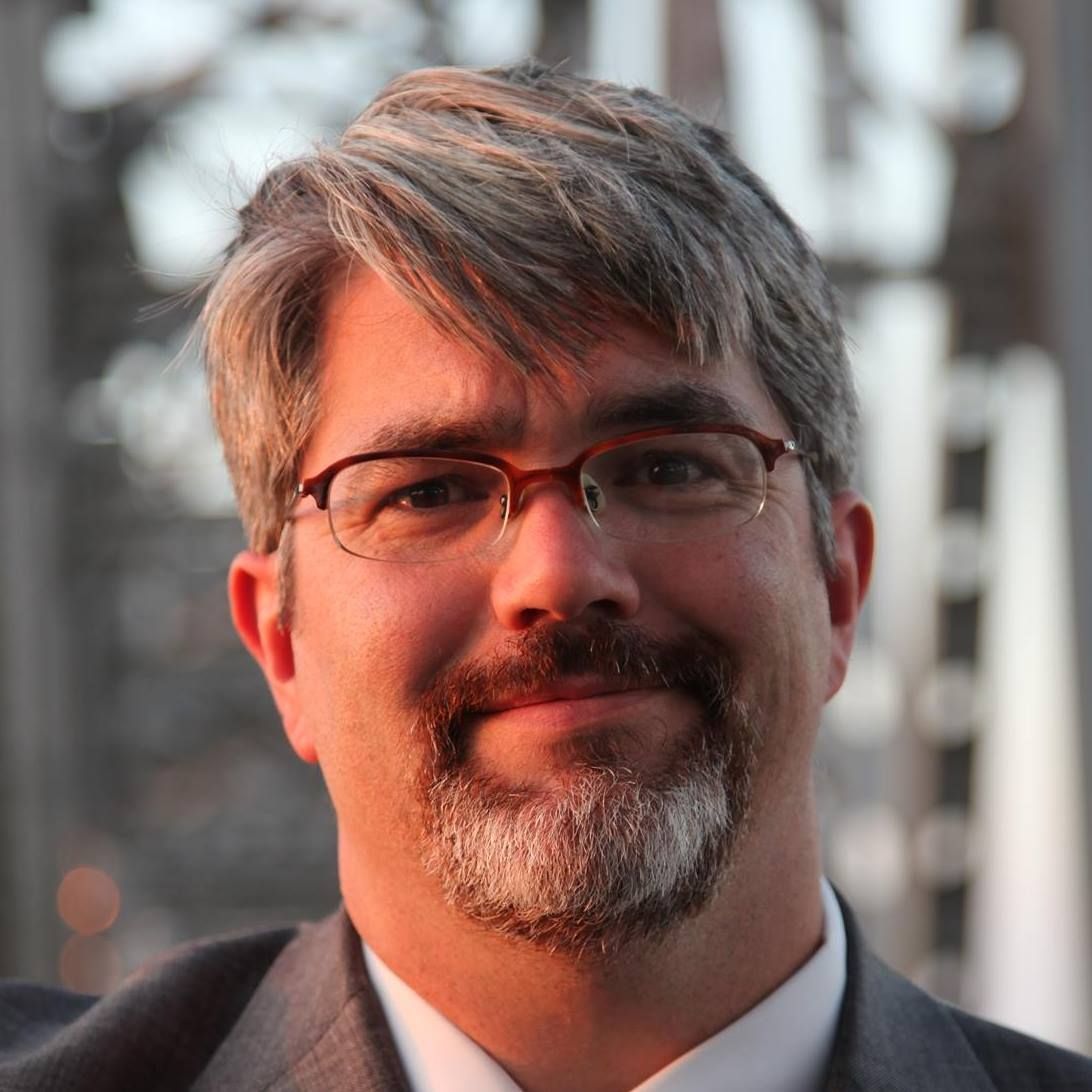 Man in suit, tie, and glasses leans against a metal bridge, smiling at the camera.