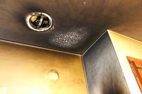 A ceiling and wall corner with dark smoke staining, a recessed light fixture, and a patch of white powdery residue.