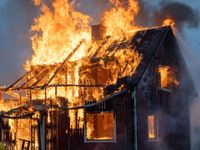 A wooden house engulfed in bright orange flames and smoke, with the roof structure partially collapsed.