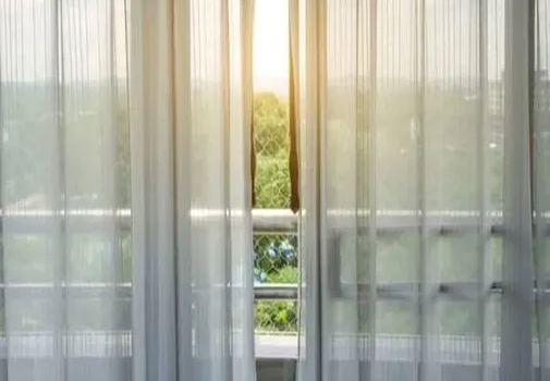 Inside view from first floor balcony, looking out through slightly open sheer blinds, with green foliage and the sunset in the background.