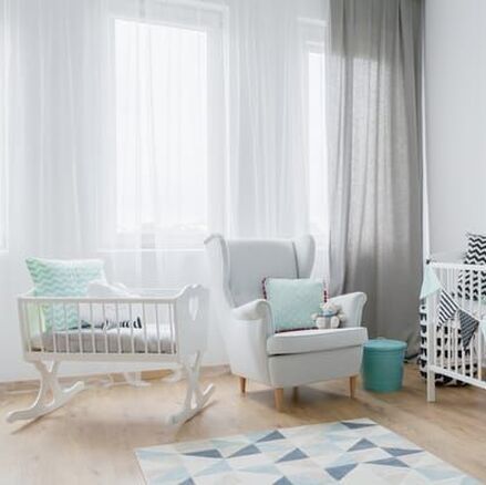 Baby nursery with timber floor, white baby's cot, white chair with soft blue cushion and stuffed toy, multi-coloured mat on the floor. Sunlight pouring in tall windows covered with sheet white curtains, framed by grey sheer curtains.