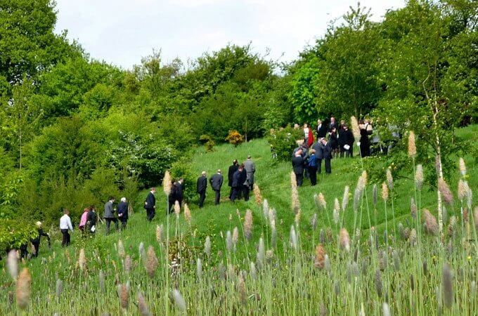 People in dark clothing walking uphill on a grassy hillside towards trees, likely a funeral or memorial.