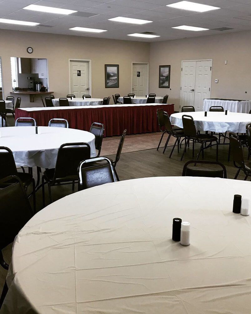 Large room set up for an event with round tables covered in white tablecloths. Brown chairs and neutral walls.