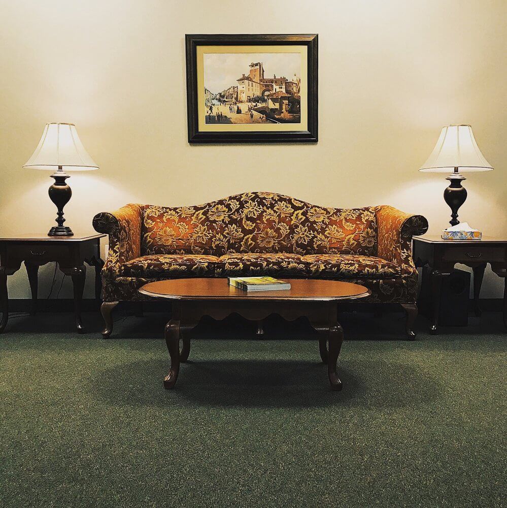 A patterned couch, side tables with lamps, coffee table, artwork on the wall, and carpet flooring.