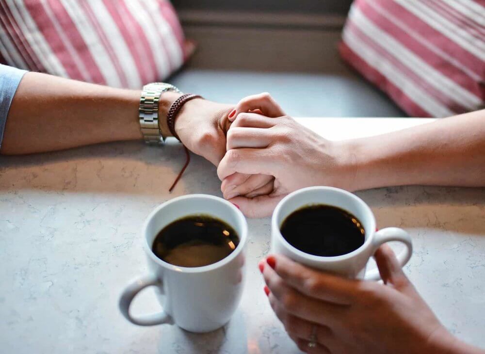Two people holding hands over coffee cups on a table, with a striped pillow in the background.