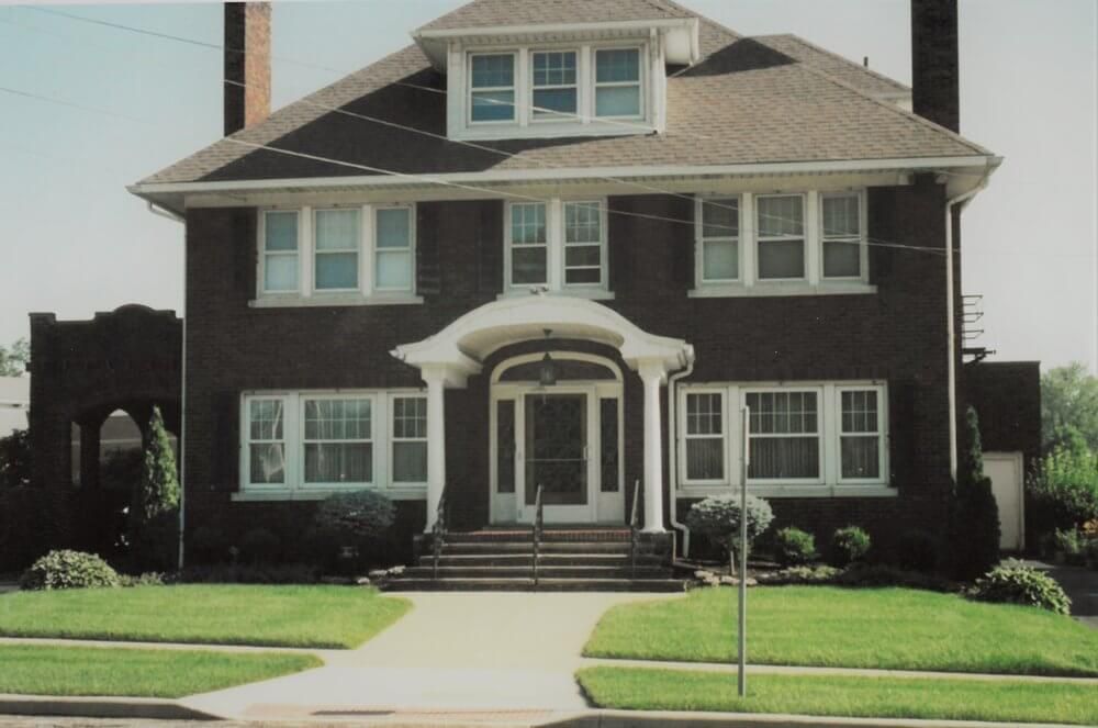Two-story brick house with white trim, steps, and covered entryway. Green lawn and sidewalk in front.