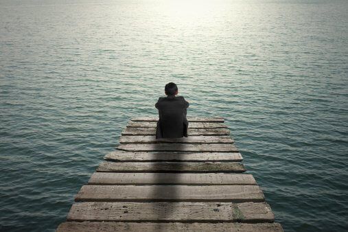 Person sits alone on a wooden dock, facing calm water.