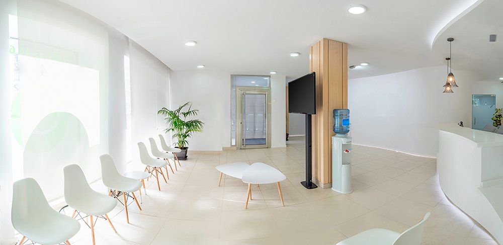 A waiting room with white chairs , a table , a television and a water cooler.