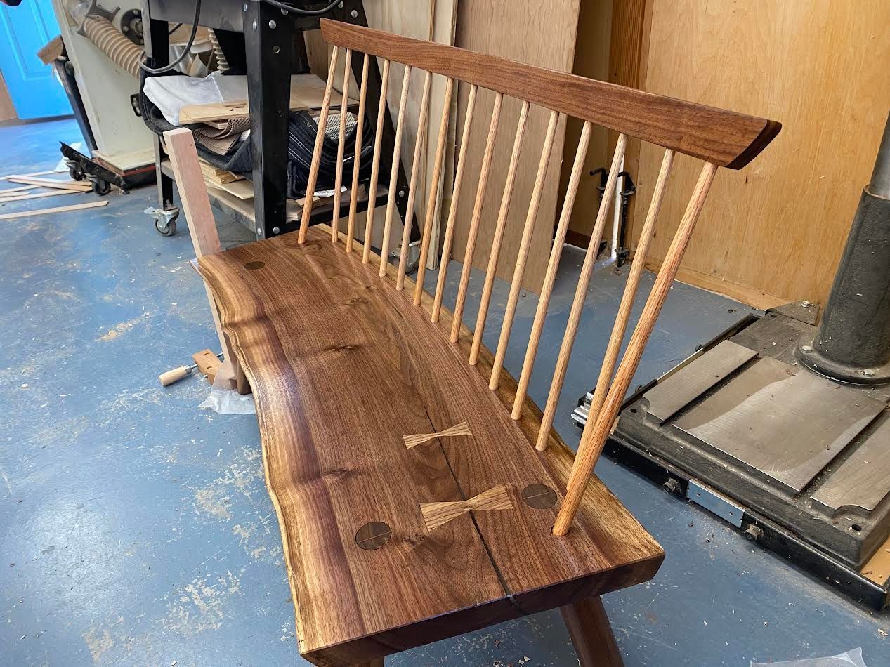 Wooden bench with live-edge seat, light spindles, and dark walnut backrest, in a workshop setting.