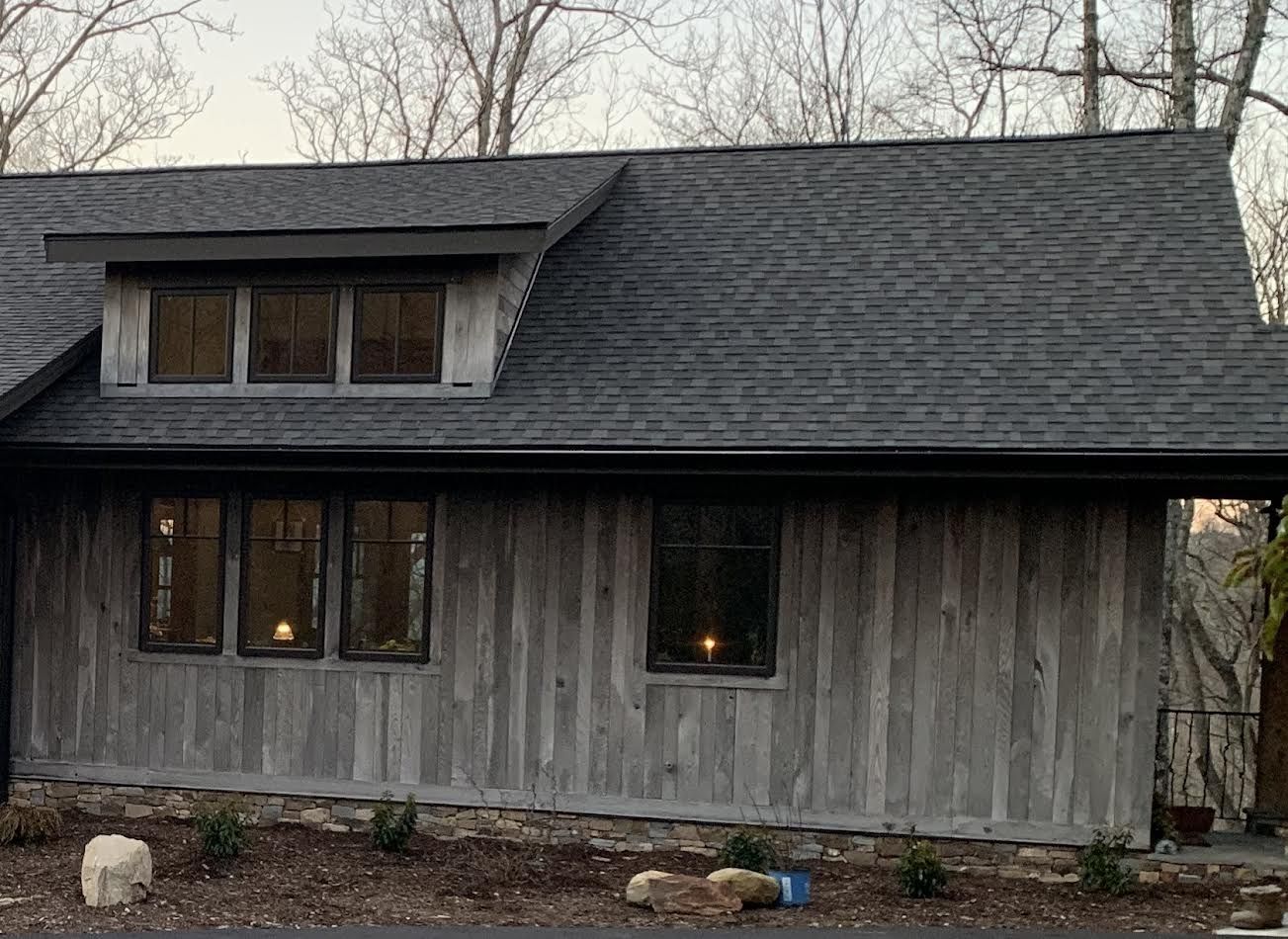 A rustic gray cabin with dark gray roof, windows, and weathered wooden siding, in a wooded setting.