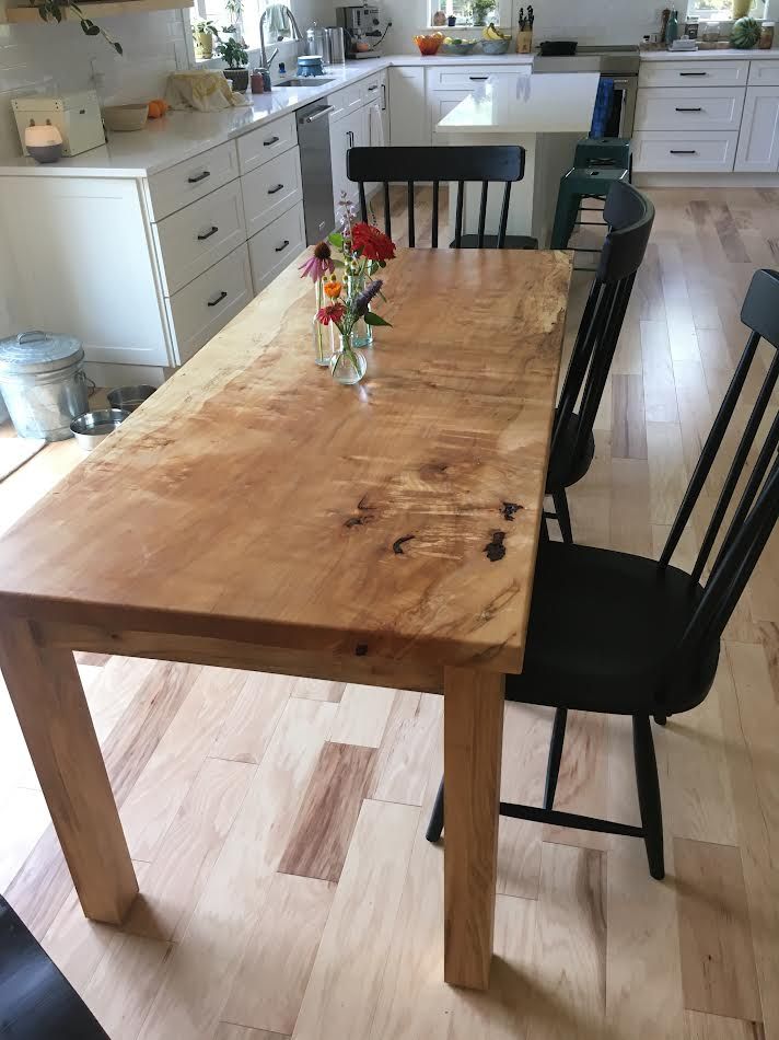 Wooden dining table with black chairs in a bright kitchen. Flowers in small vases.