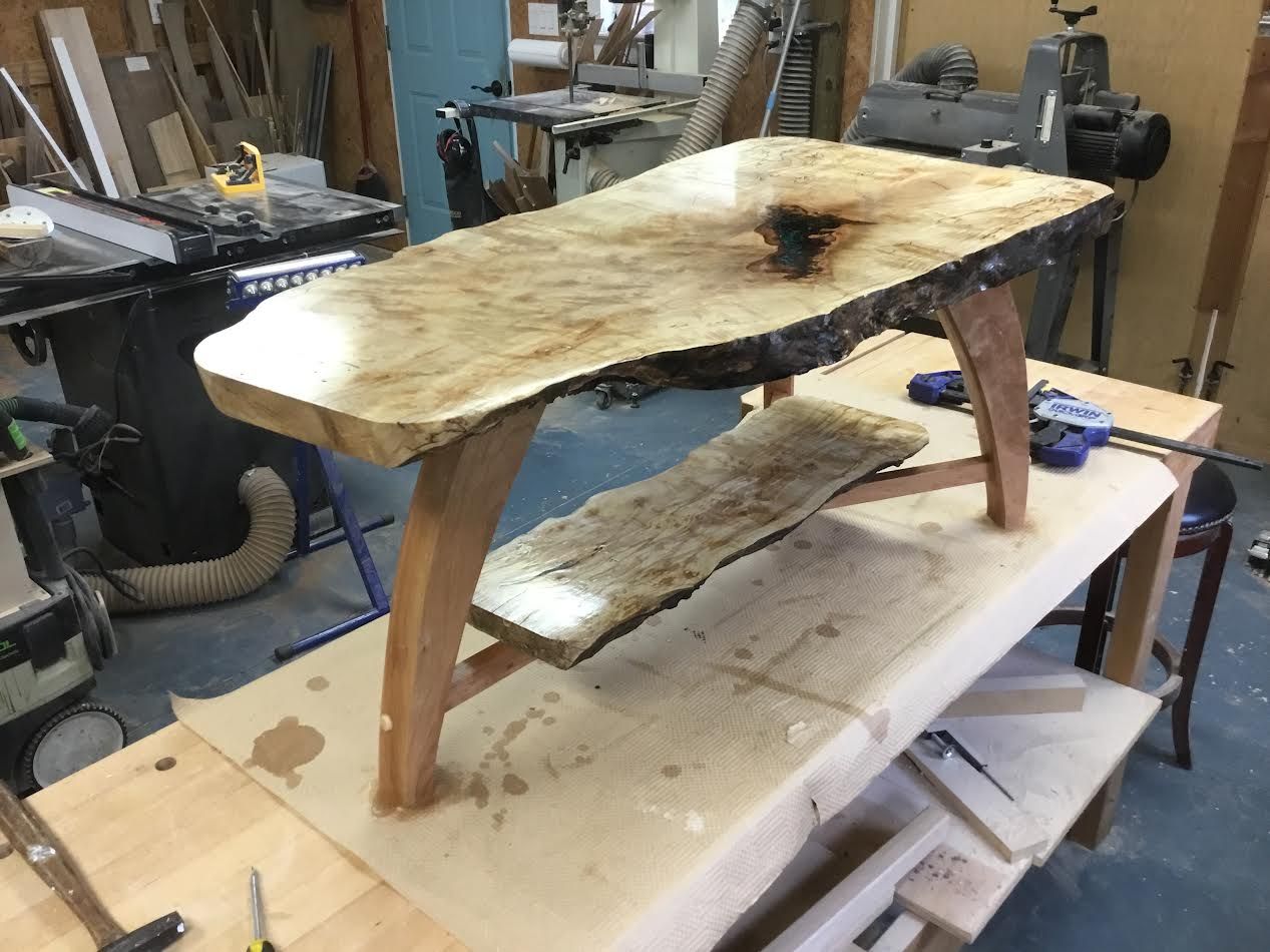 Wooden table with live edge top and lower shelf, sitting on a work bench in a workshop.