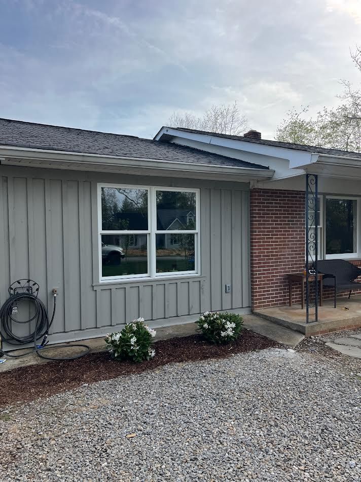 Gray house with white-framed window. Brown mulch and gravel landscaping in front. Cloudy sky.