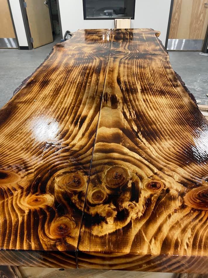 Close-up of a large, shiny wooden table. The wood has a scorched, textured appearance with visible knots and grain.