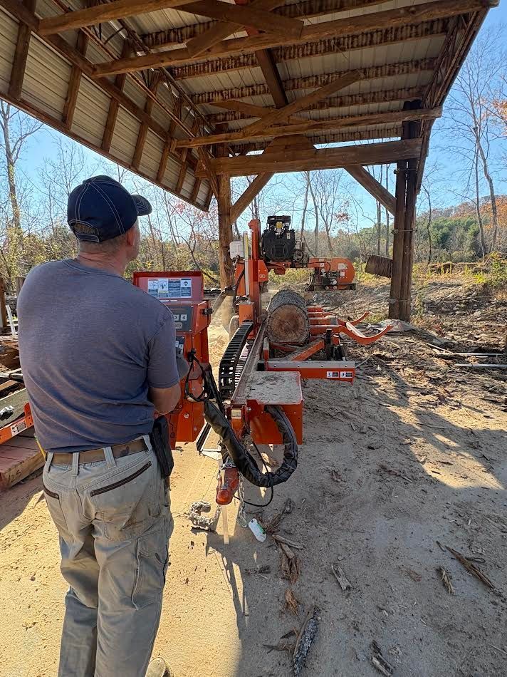 Man operating a sawmill under a wooden shelter, sawing a log outdoors.