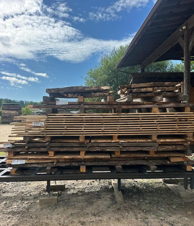 Stacks of rough-cut lumber on pallets outdoors under a blue sky, near a building.