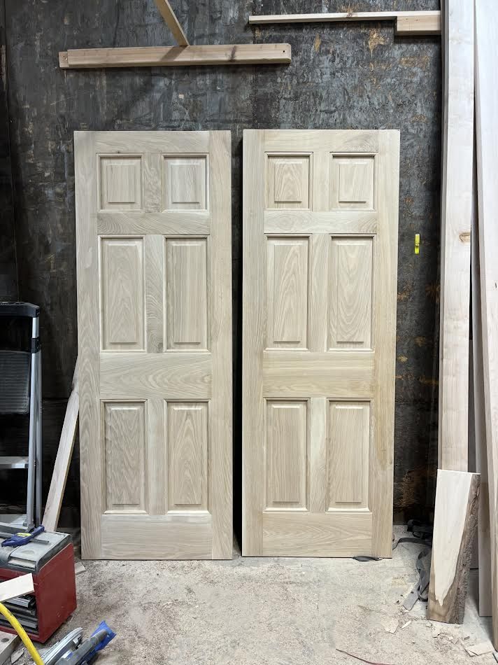 Two unfinished, six-panel wooden doors leaning against a wall in a workshop.
