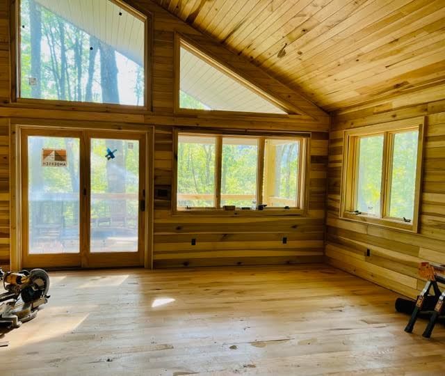 Interior view of a wooden room under construction with windows and a doorway looking out into a forest.