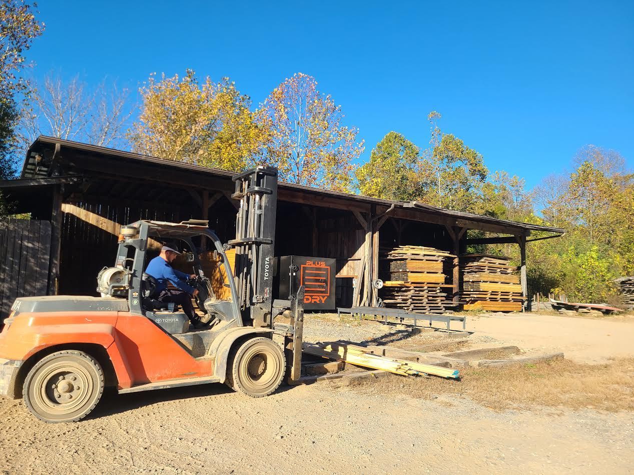 Forklift unloading lumber from a wooden shelter on a gravel lot. Orange and black forklift, sunny day.