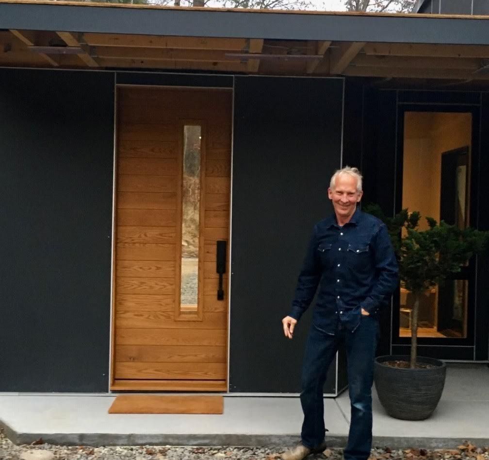 Man standing in front of a modern wooden door with a large glass panel. Black exterior, potted plant.