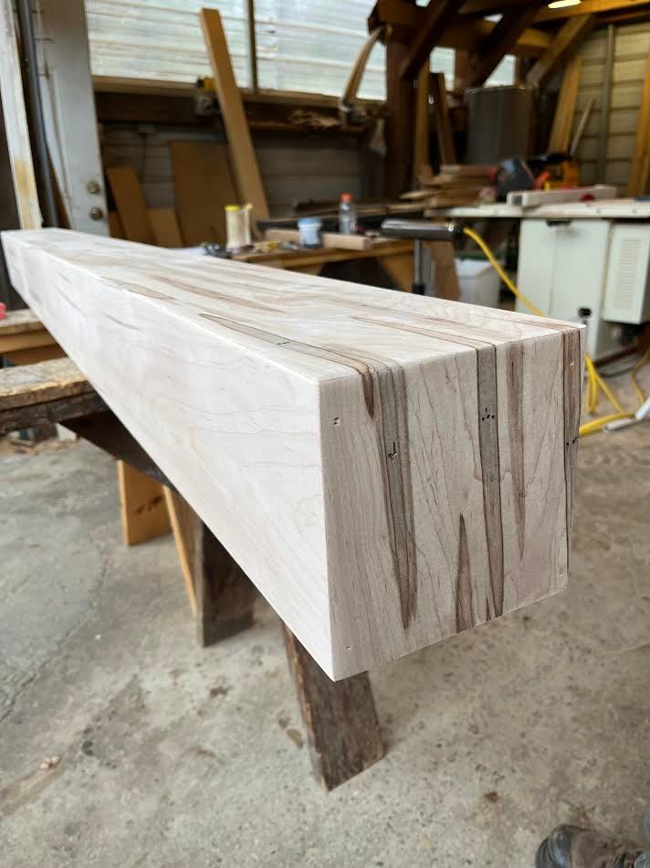 A light-colored, rectangular wooden beam with dark streaks, resting on a wooden sawhorse in a workshop.