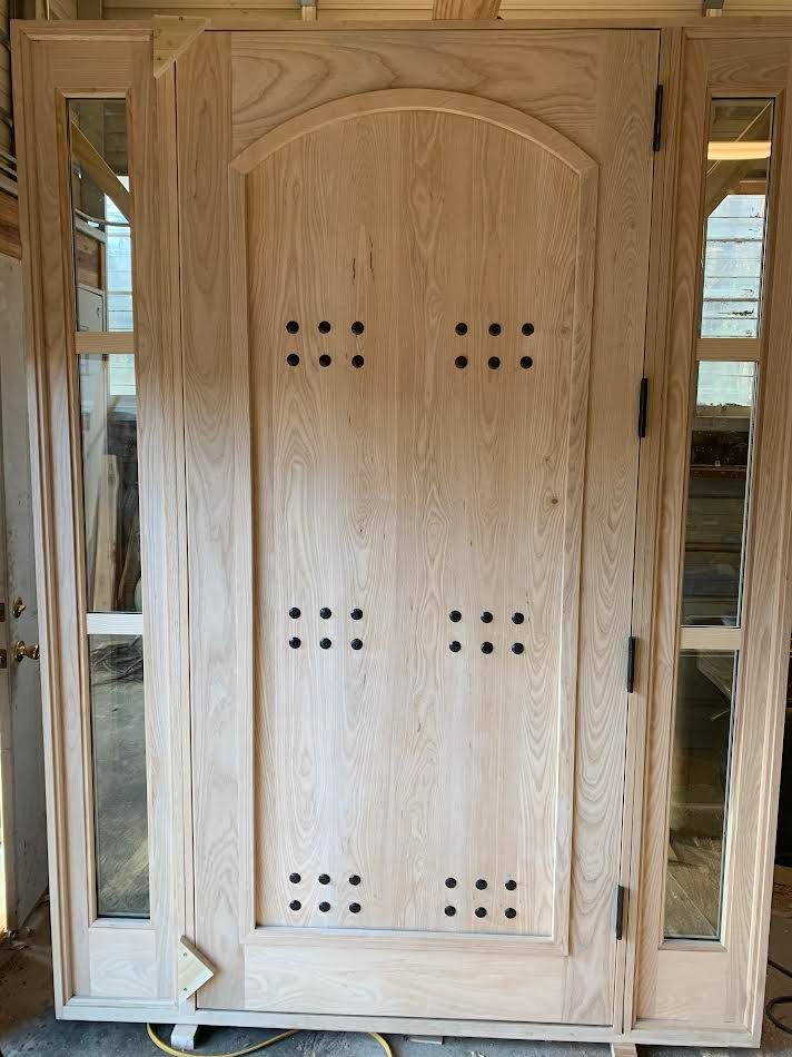 Wooden front door with sidelights, featuring decorative panels and hardware; unstained and ready for finish.