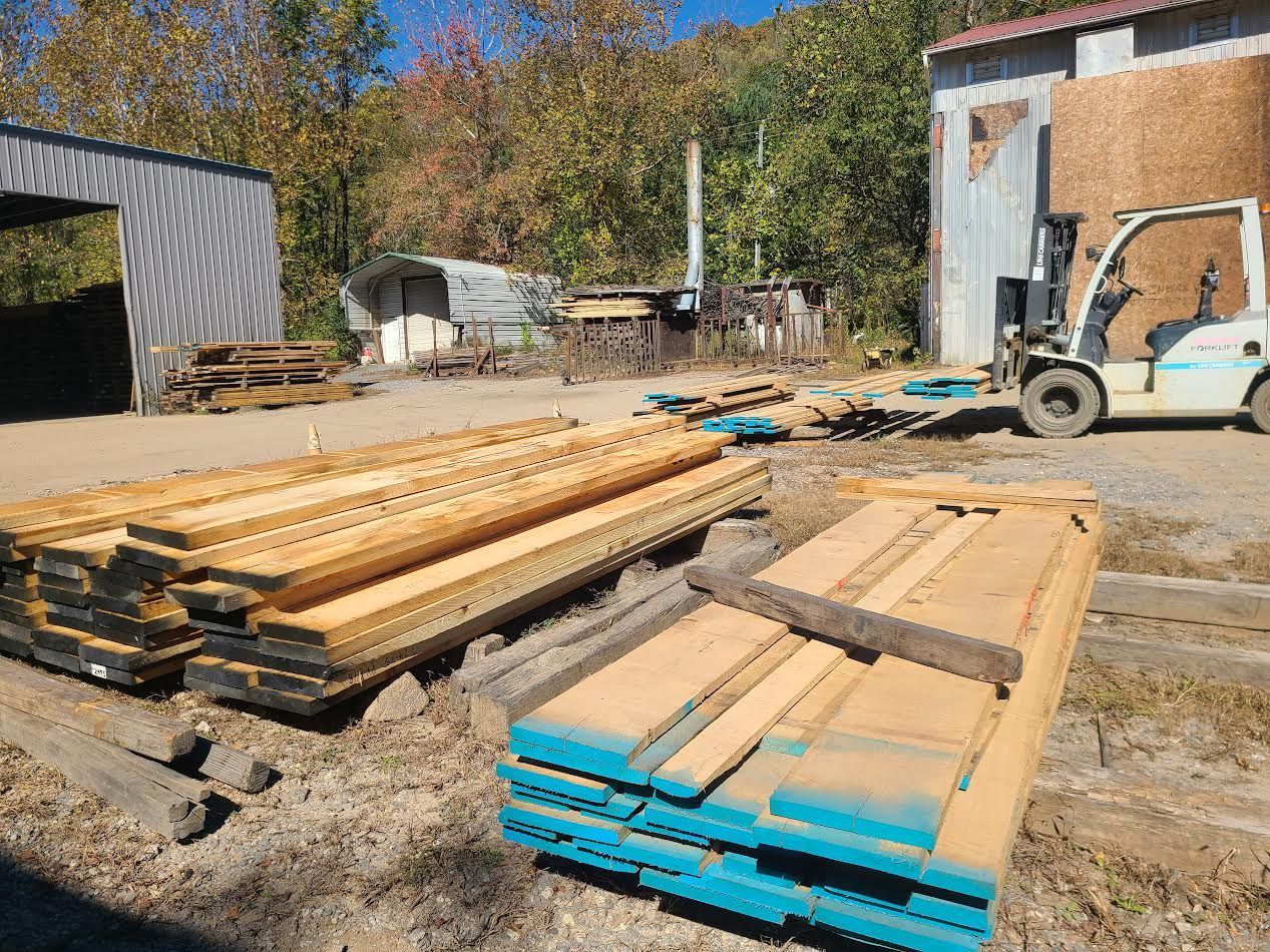 Lumberyard with stacked wood, forklift, and sheds on a sunny day. Blue paint on wood ends.
