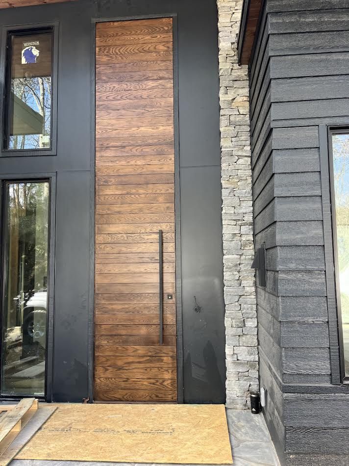 Exterior of a modern home with a large wooden door and black metal siding. Stone accent pillar on the right.
