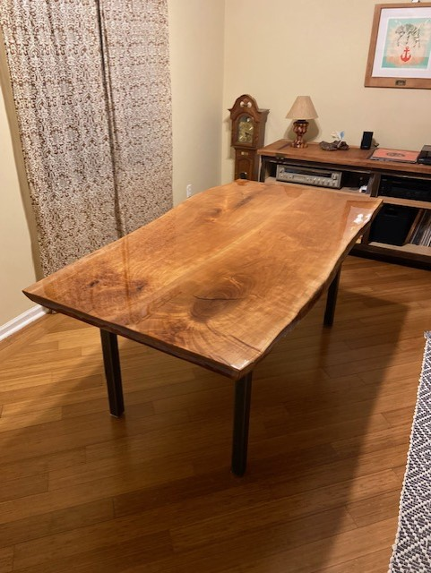 A wooden table with live edge, brown wood, black legs, in a room with hardwood floors, a curtain, and a clock.