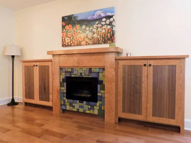 Wooden fireplace with colorful tile surround, flanked by matching cabinets, topped with floral artwork.