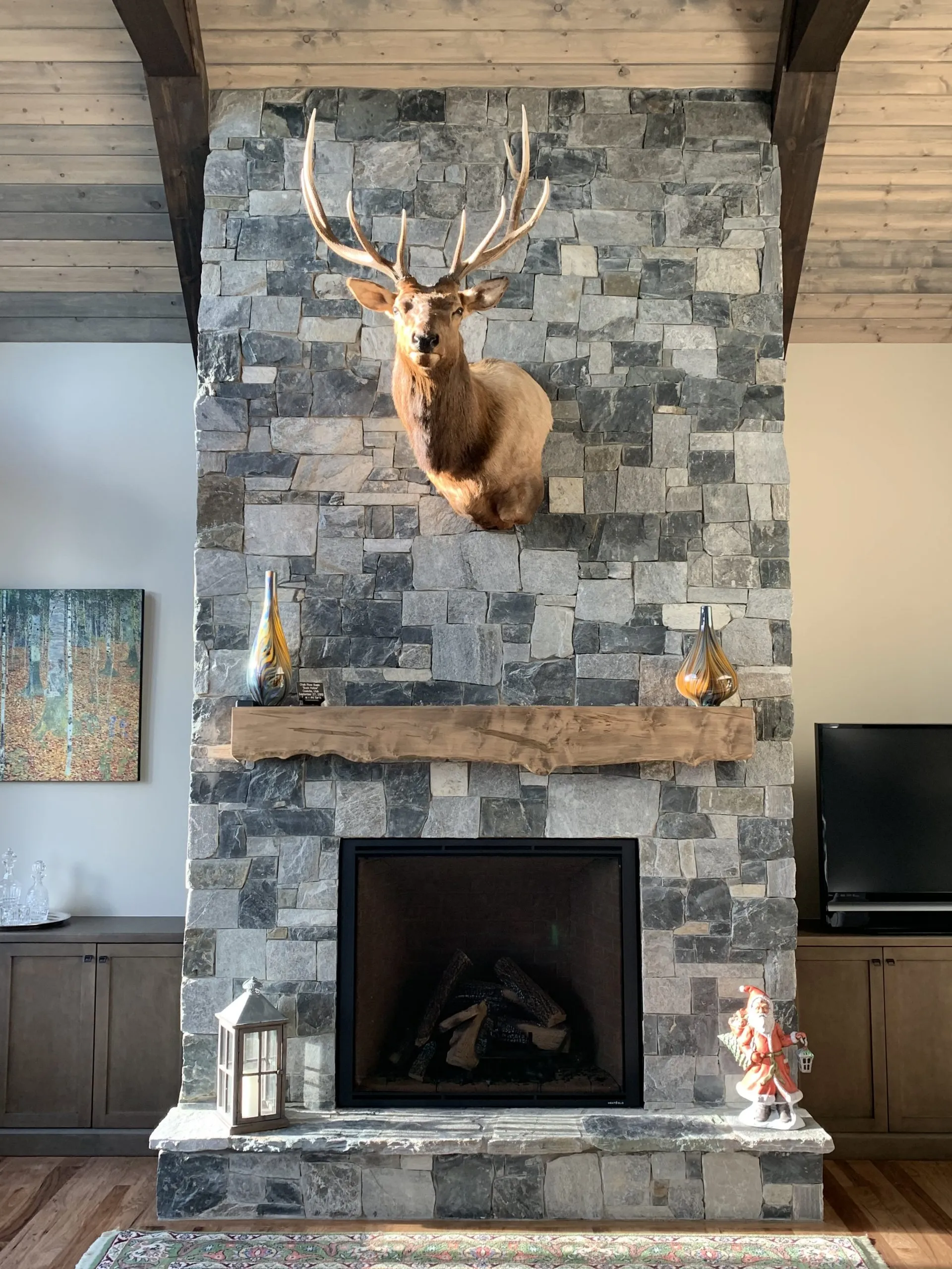 Fireplace with stone facade, elk head mount, wooden beam mantel, and cabinets.