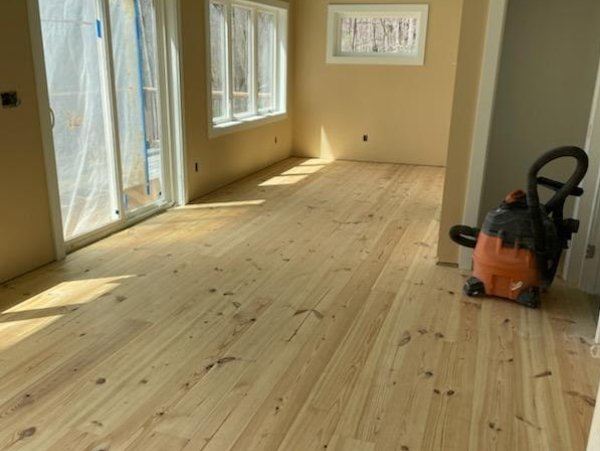 Newly installed light-colored wood floor in a room with windows and an orange vacuum cleaner.