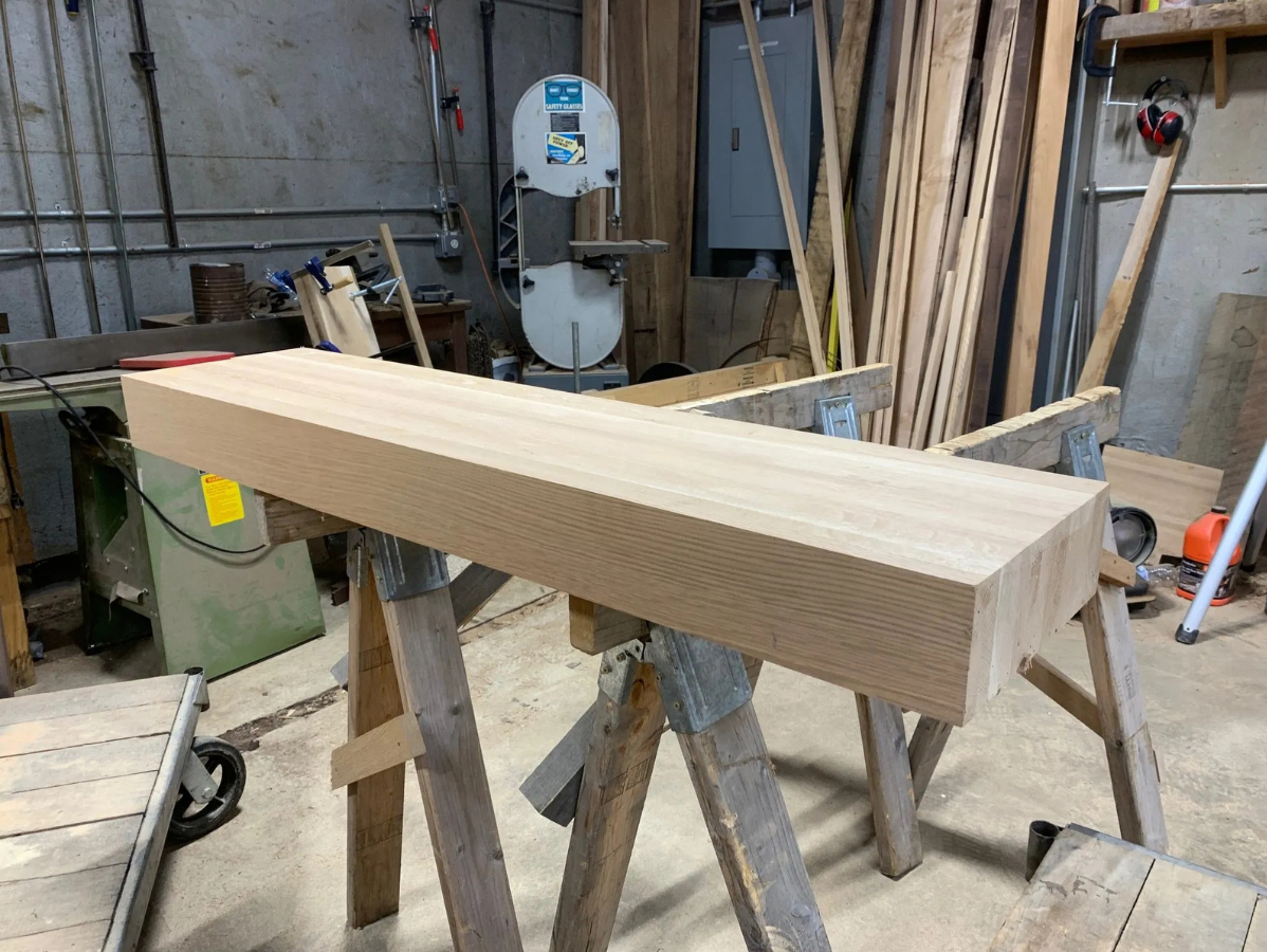 Wooden beam on sawhorses in a workshop with a bandsaw and other woodworking tools in the background.