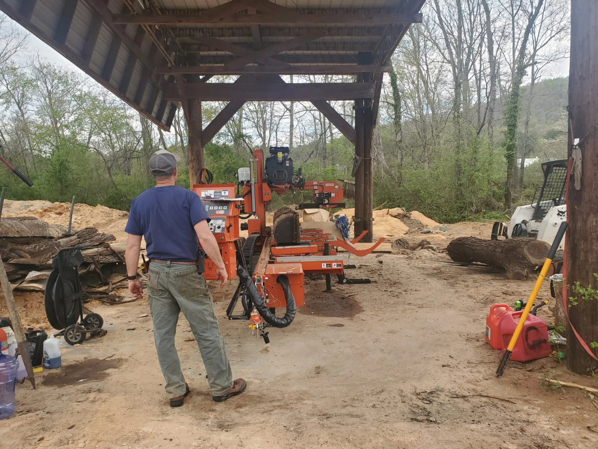 Man operating an orange sawmill under a wooden shelter, with logs and equipment nearby.