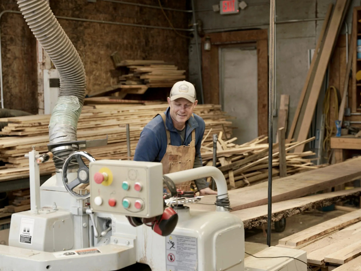 Woodworker in a workshop, smiling. Operating a machine with wood pieces in the background.