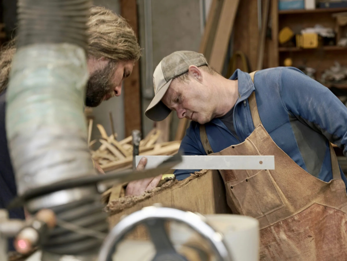 Two people working in a wood workshop, one using a tool on a wooden object, the other watching.