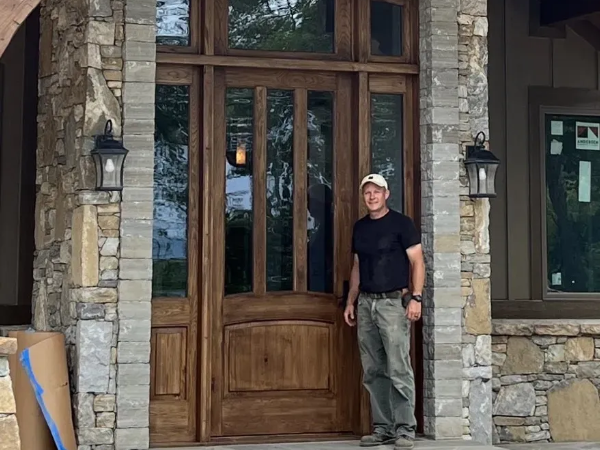 Man standing in front of a wooden door with sidelights, flanked by stone columns; exterior.