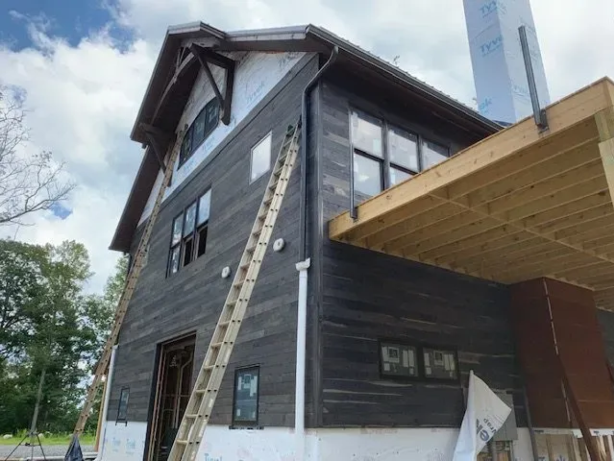Partially constructed house with dark siding, windows, ladder, and unfinished deck.